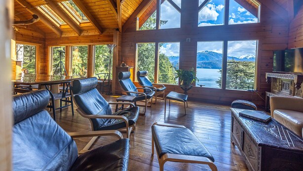 A wooden cabin with large windows offering mountain views, leather seating, and a wooden dining table.
