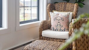 A wicker chair with a cushion and a patterned pillow, positioned near a window with white curtains. - 738 Alice Loop - 4 Sea's Cottage (Sitka)