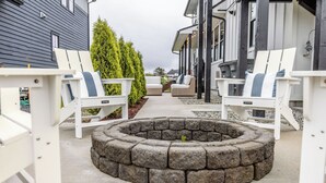 A fire pit surrounded by white Adirondack chairs, a stone fire pit, and a pathway leading to a building with a dark exterior and white trim. - 738 Alice Loop - 4 Sea's Cottage (Sitka)