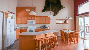 Een moderne keuken met houten kastjes, een centraal eiland met krukken, een berenbeeld aan de wand en een groot raam.