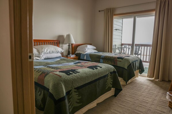 A hotel room with two beds, a nightstand with a lamp, and a balcony with a table and chairs. - Chichigof Suite at Island View (Sitka)