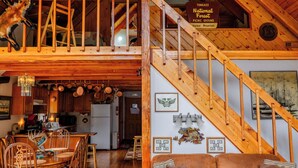 Un vaste espace de vie de style loft, doté d’un plafond haut, de poutres apparentes, d’un coin repas avec une table et des chaises en bois, d’un canapé en cuir et d’un escalier avec une rampe en bois.