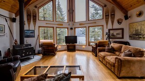 Un salon spacieux doté d’une grande cheminée, de parquets en bois et de grandes fenêtres offrant une vue sur les arbres.