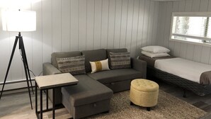 Habitació moderna i ben il·luminada amb un sofà gris, una taula petita, una làmpada de peu, un llit i una finestra amb persianes.