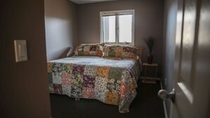 Un dormitorio con una cama cubierta con un colcha de retazos, una ventana con persianas y una puerta con pomo.