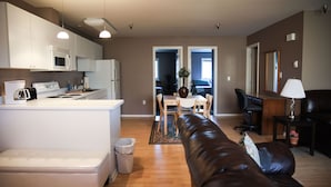 A modern kitchen with white appliances, a dining area with a table and chairs, a leather sofa, and a small side table with a lamp.