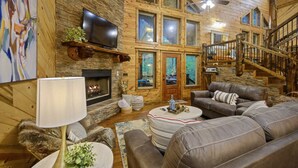 Un acogedor interior de cabaña con chimenea, un televisor montado en una pared de piedra, una zona de estar confortable con sofá y sillones, y una escalera que conduce a un nivel superior.