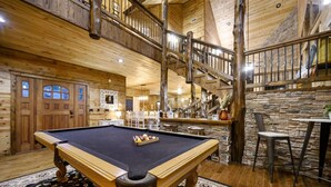 Una mesa de billar en una sala con piso de madera, una escalera y una pared de piedra.