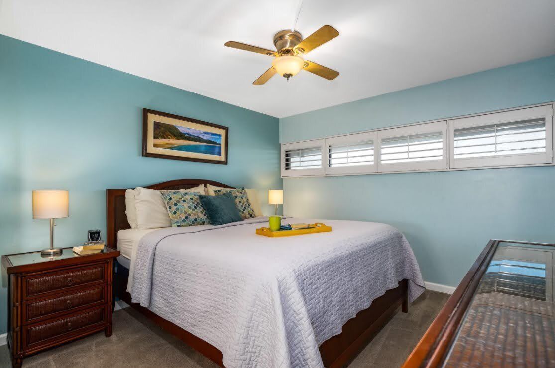 Une chambre à coucher comprenant un lit, des tables de chevet, un ventilateur de plafond, une fenêtre avec des stores et un tableau accroché au mur.