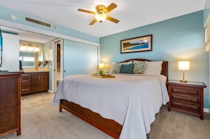 A bedroom with a bed, bedside tables, a ceiling fan, a painting, and a bathroom with a mirror and sink.