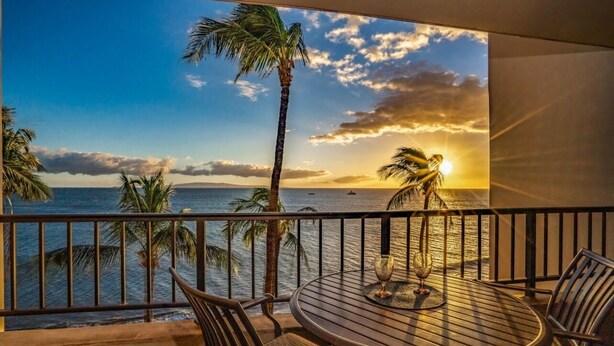 Une table ronde entourée de chaises, sur un balcon offrant une vue imprenable sur le coucher de soleil, avec des palmiers et l’océan en toile de fond.
