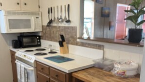 Una cocina compacta con armarios blancos, microondas, estufa y fregadero. Hay un cubo de basura y una planta en maceta sobre la encimera.
