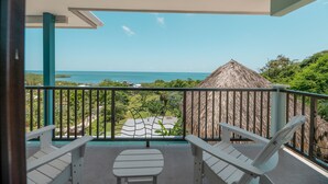 A balcony with two white chairs and a small table, overlooking a beach with a thatched-roof hut and lush greenery. - Sweeping panoramic views of the reef  (Bay Islands Department)