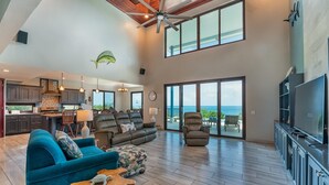 A spacious living area with a large sofa, a coffee table, and a television. There is a kitchen area with cabinets and a dining table. The room has large windows and a ceiling fan. - Sweeping panoramic views of the reef  (Bay Islands Department)