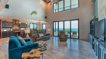 A spacious living area with a large sofa, a coffee table, and a television. There is a kitchen area with cabinets and a dining table. The room has large windows and a ceiling fan.