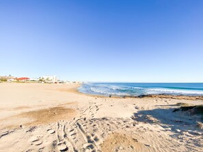 A beach with footprints leading to the water, buildings in the background, and a clear sky. - PINK SEAFRONT (Torrevieja)