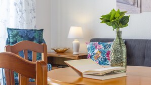 A wooden table with an open book, a glass vase with green plants, a lamp, and a colorful pillow on a couch. - PINK SEAFRONT (Torrevieja)