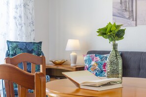 Uma mesa de madeira com um livro aberto, um vaso de vidro com plantas verdes, uma lâmpada e uma almofada colorida em um sofá.