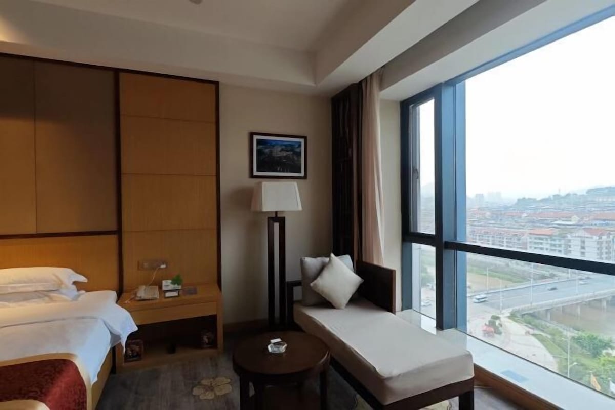 Habitación de hotel con cama, sofá, una mesita, lámpara y un ventanal con vistas a la ciudad.