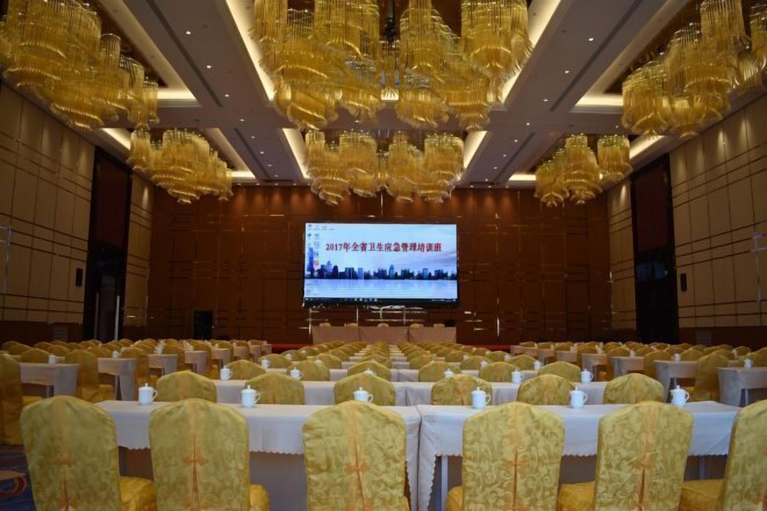 Un salón de conferencias con filas de mesas y sillas, una pantalla grande al frente y candelabros.