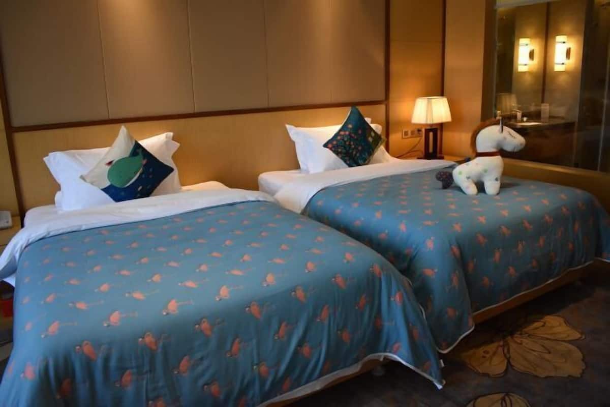 A hotel room with two beds, each with a blue bedspread and white pillows. There is a lamp on the nightstand and a stuffed toy on the bed.