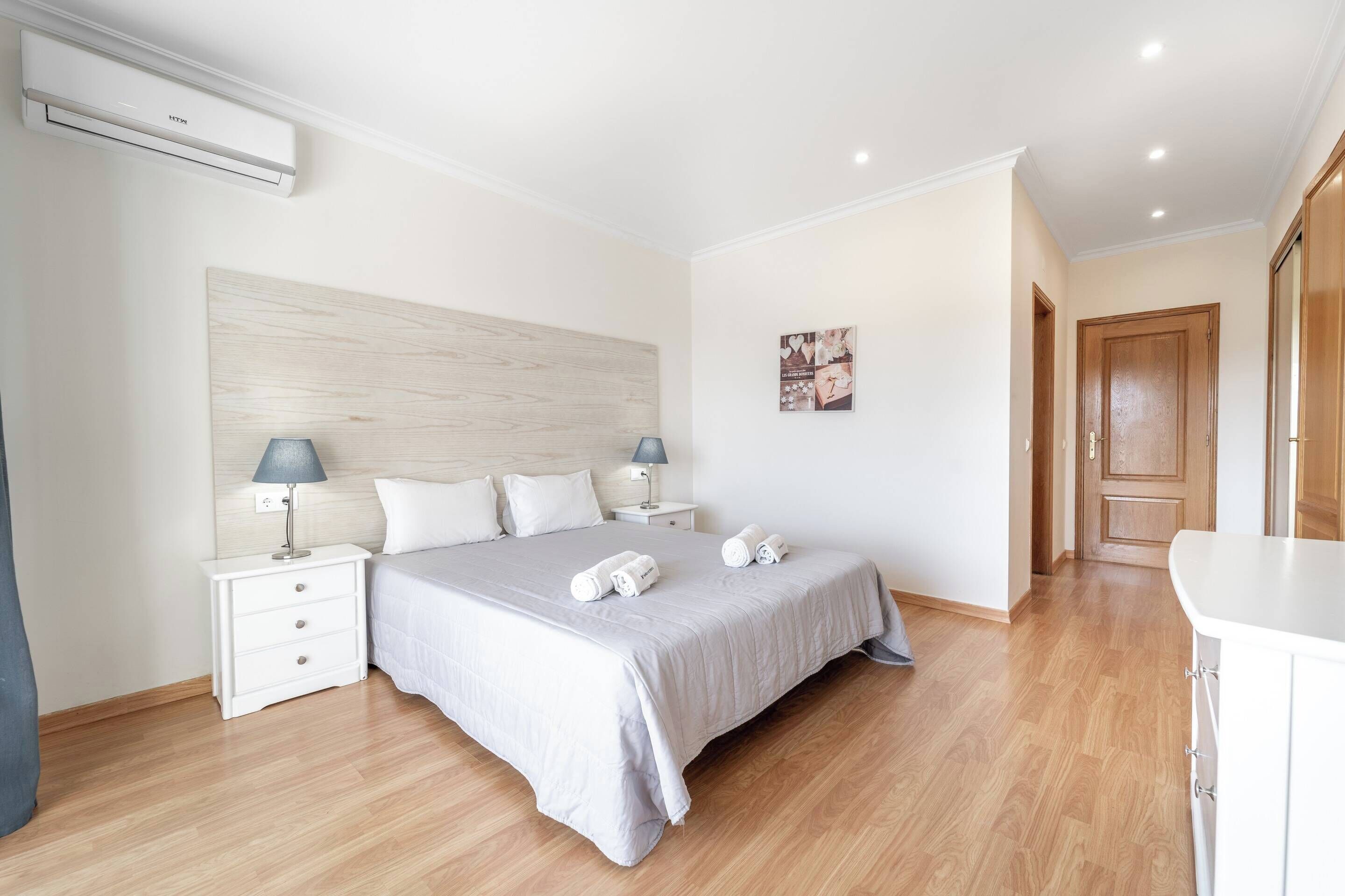 A modern bedroom with a large bed, two bedside tables with lamps, a wooden headboard, and a wooden floor.