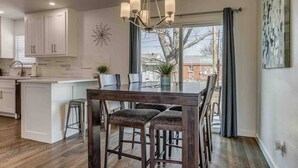 A modern kitchen with a central island, a dining area with a dark wood table and cushioned chairs, and a large window with a view of the outside. - Luxury Modern 3BR - Private Primary Suite! (Oklahoma City)