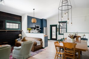 A modern kitchen with dark blue cabinets, a hanging light fixture, and a dining area with a wooden table and chairs.