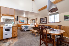 A cozy room with a bed, a wooden dining table, and a kitchen area with a stove and cabinets. - Ravens Heart Cabin (West Yellowstone)