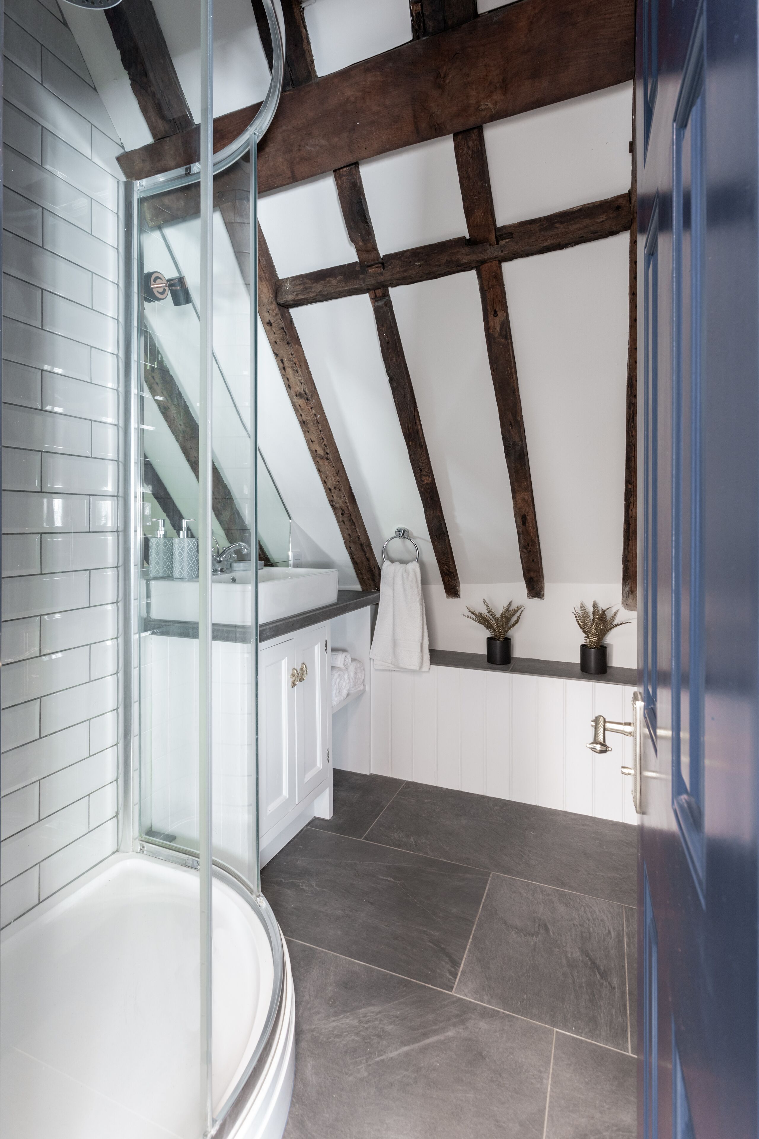 A modern bathroom with a glass shower enclosure, white tiled walls, dark stone flooring, and a wooden ceiling beam.