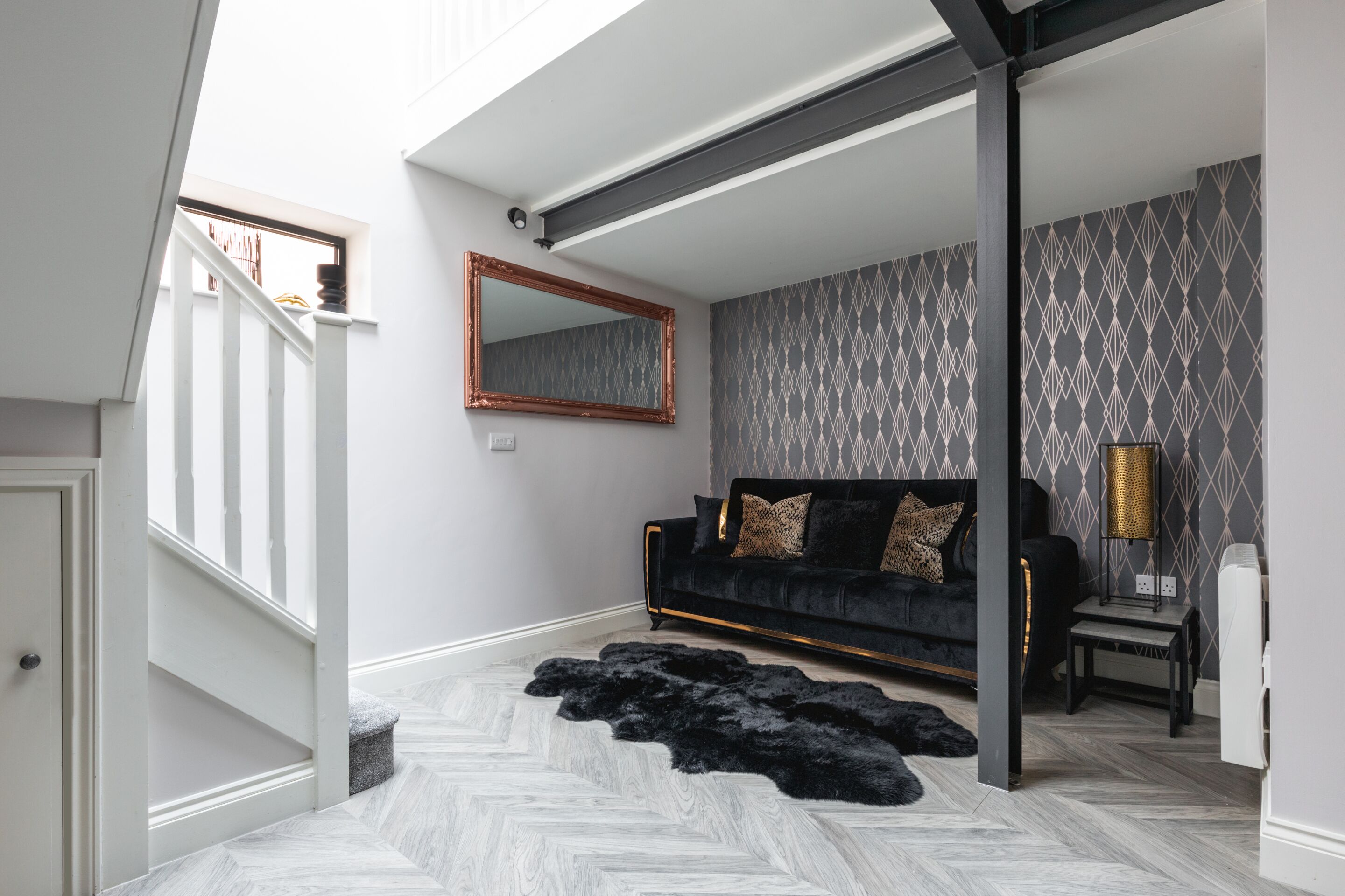 A modern living room with a black sofa, patterned wallpaper, and a herringbone floor.