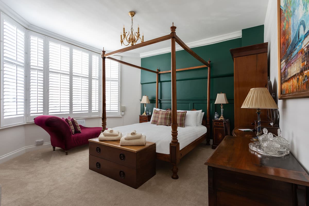 A four-poster bed with a wooden frame, a red upholstered chair, a wooden nightstand with a lamp, and a painting on the wall.