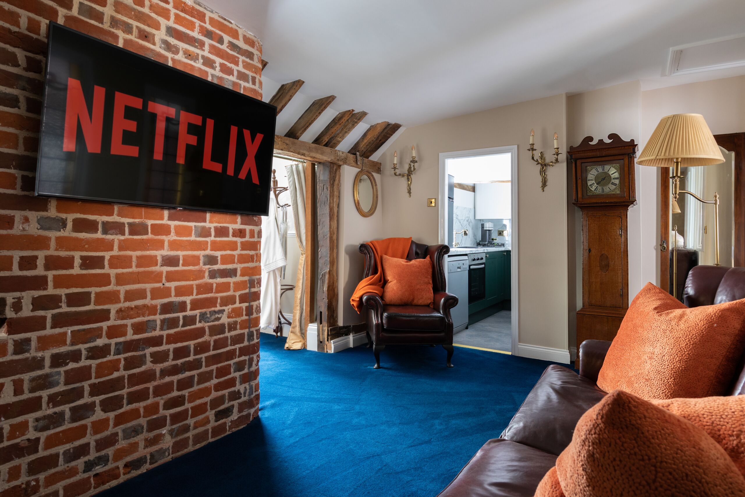 A living room with a brick wall, a Netflix sign, a brown leather sofa, an orange armchair, and a blue carpet.