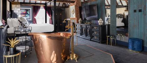 A modern room with a large copper bathtub, a bed, a TV, and a dining area with stools.