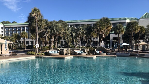 A pool area with lounge chairs and umbrellas, surrounded by palm trees and a building with a green roof. -  Poolside #, Westin Amenities Included with your stay.  #133 (Hilton Head Island)