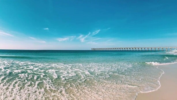 A beach with clear turquoise water, a sandy shore, and a pier extending into the sea.