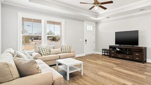 A living room with a beige sofa, a white coffee table, a TV on a wooden stand, and large windows with blinds.