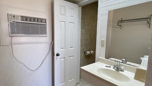 Un bagno con lavandino bianco, specchio e porta asciugamani. Si vede una porta con un asciugamano appeso e un cestino.