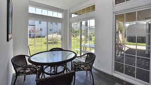 A dining area with a round glass table and wicker chairs, large windows offering views of a garden and a barn. - Book Now for Fun  in the Sun! Spring  & Summer 2026  (Narragansett)