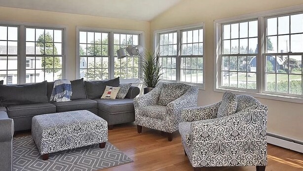 A spacious living room with a sectional sofa, two patterned armchairs, and a matching ottoman. Large windows provide ample natural light and a view of the outdoors. - Book Now for Fun  in the Sun! Spring  & Summer 2026  (Narragansett)
