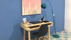 A room with a blue wall, a wooden desk, a black chair, a teal desk lamp, and a white air conditioner. - Casa 66 (Mérida)
