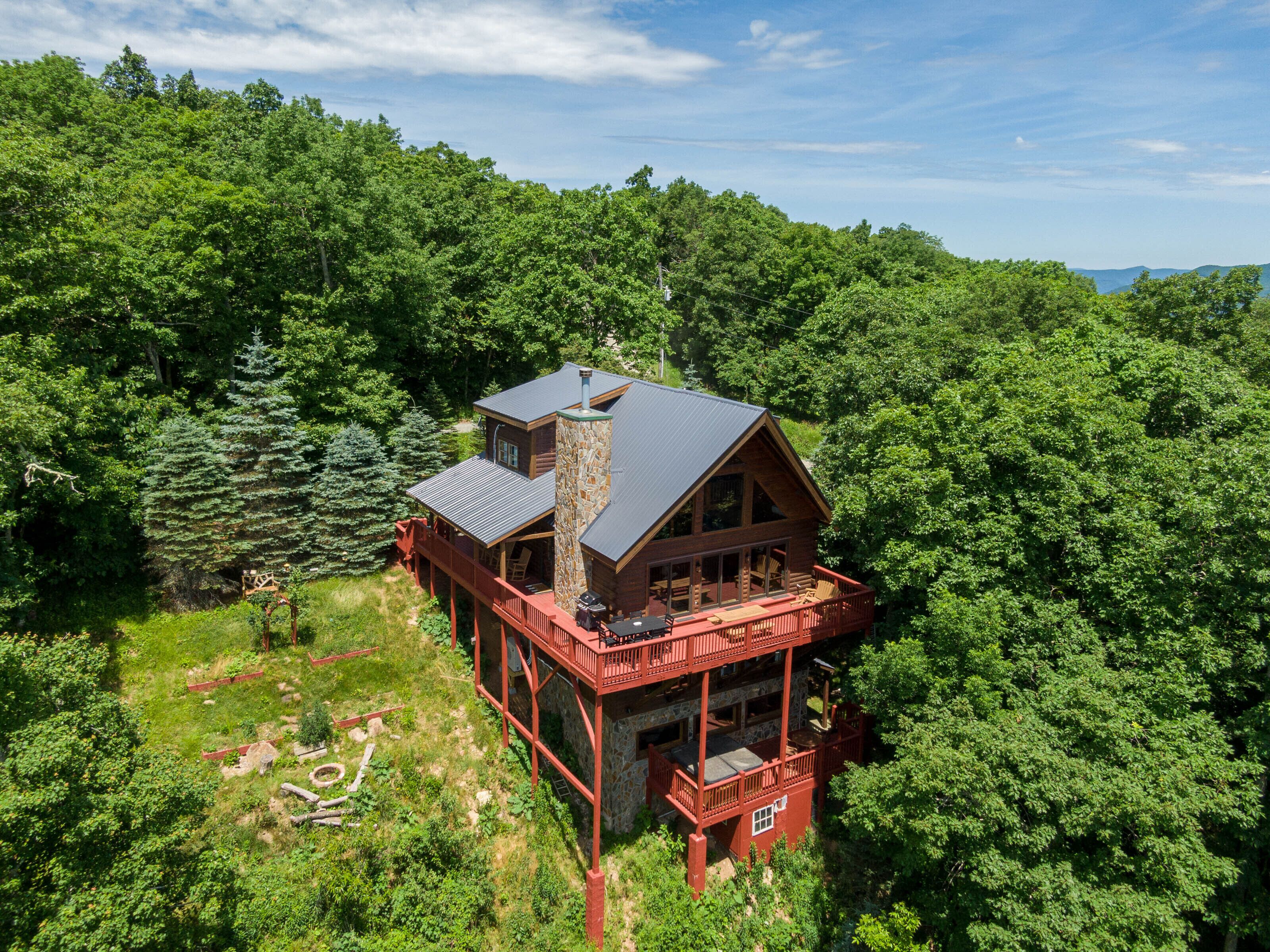 Une cabane en rondins avec une cheminée en pierre, plusieurs terrasses et entourée d’une forêt dense.