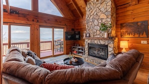A cozy cabin interior with a stone fireplace, wooden walls, a large window, and a deck with a view. - Carolina Lookout - 10 mins to downtown Boone, hot tub, ping pong, long mountain views (Boone)