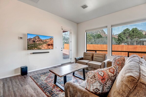 A living room with a large TV, a sofa, a coffee table, and a view of a wooden fence and trees. - Discover Sedona Grasshopper: in West Sedona with stunning red rock views. (Sedona)