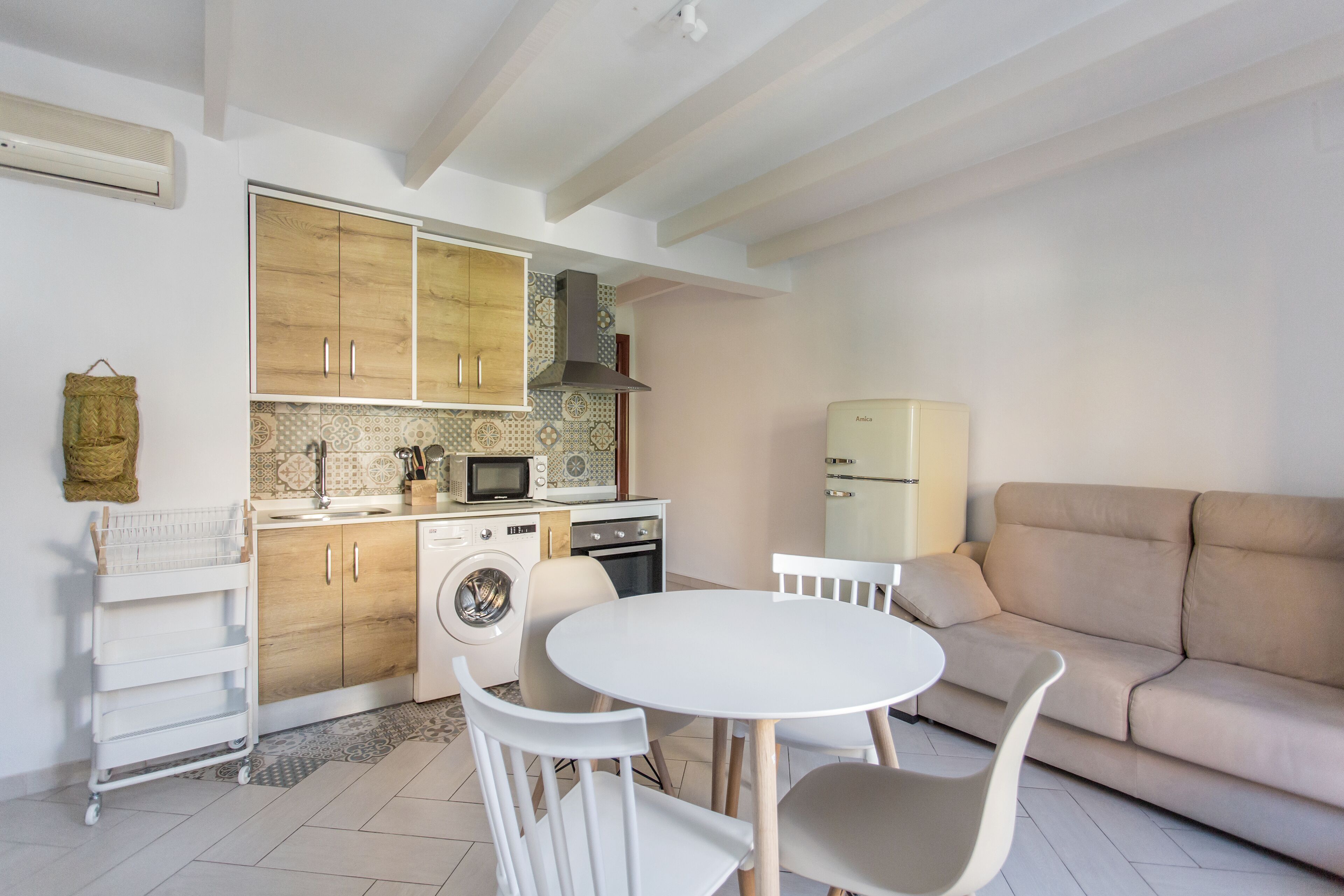 A compact living space with a kitchenette, a dining area with a round table and chairs, a washing machine, and a sofa.