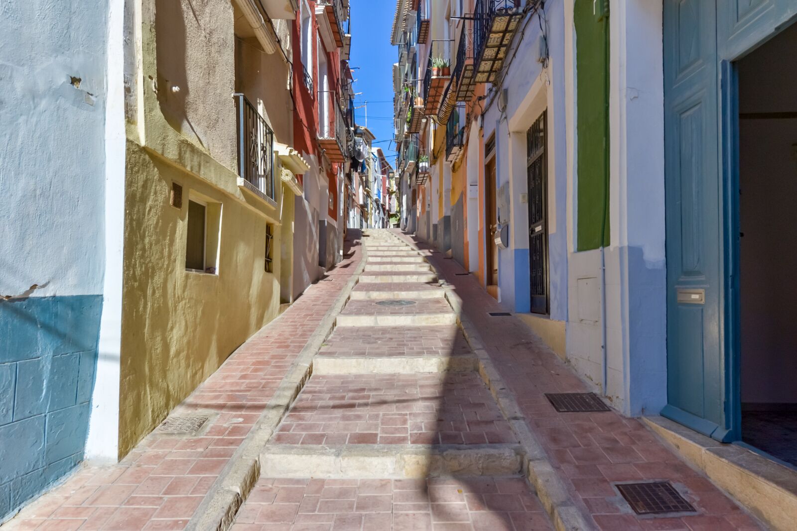 Une rue étroite et ensoleillée, bordée de bâtiments aux couleurs vives, avec un chemin pavé de briques et des balcons ornés de plantes.