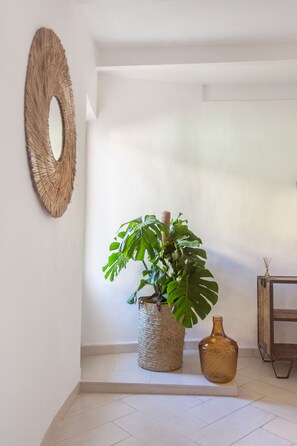 Rincón de una habitación con una planta grande en una cesta tejida, un jarrón pequeño y un espejo redondo en la pared.