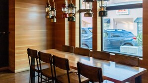 Ein Restaurant mit einem langen Holztisch und Stühlen, hängenden Pendelleuchten und Blick auf geparkte Autos durch große Fenster.