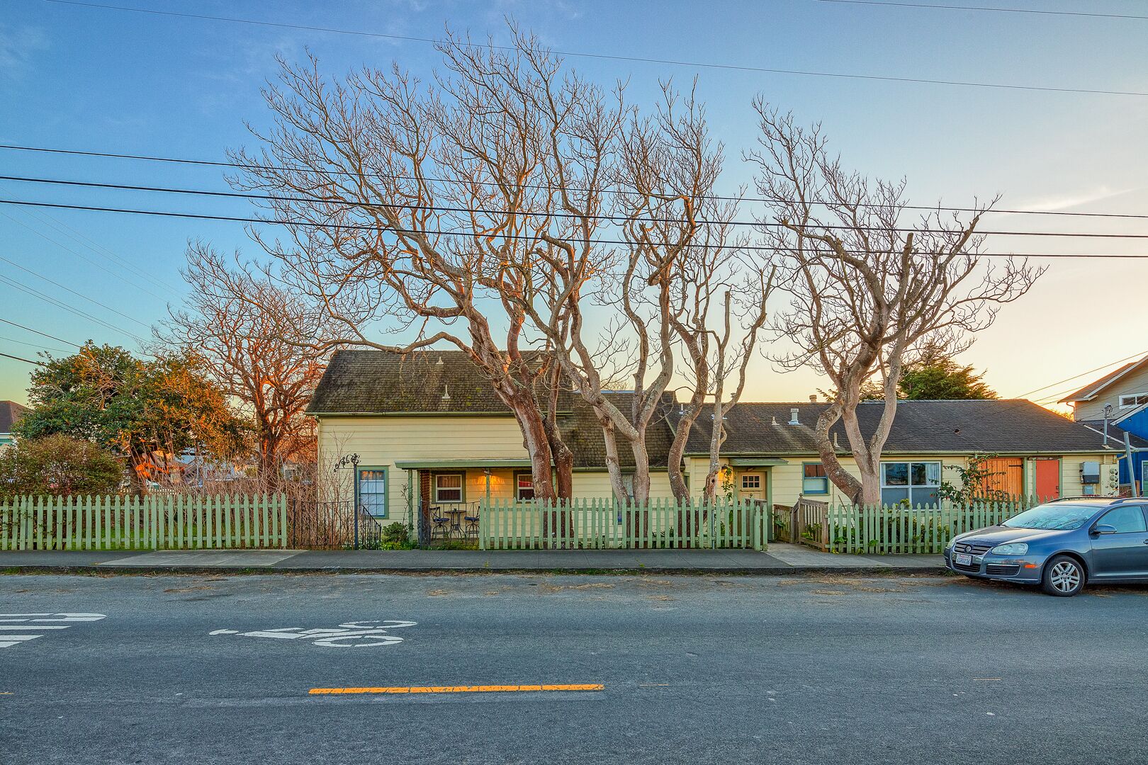 A residential street with houses, a white picket fence, and a parked car.