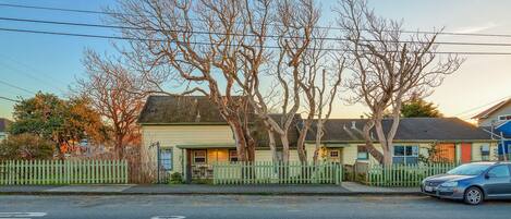 A residential street with houses, a white picket fence, and a parked car.
